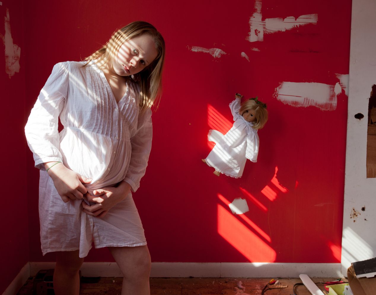 Young girl standing in a red room, buttoning her white dress, environmental portrait photography, Ilona Szwarc, contemporary Los Angeles artist.