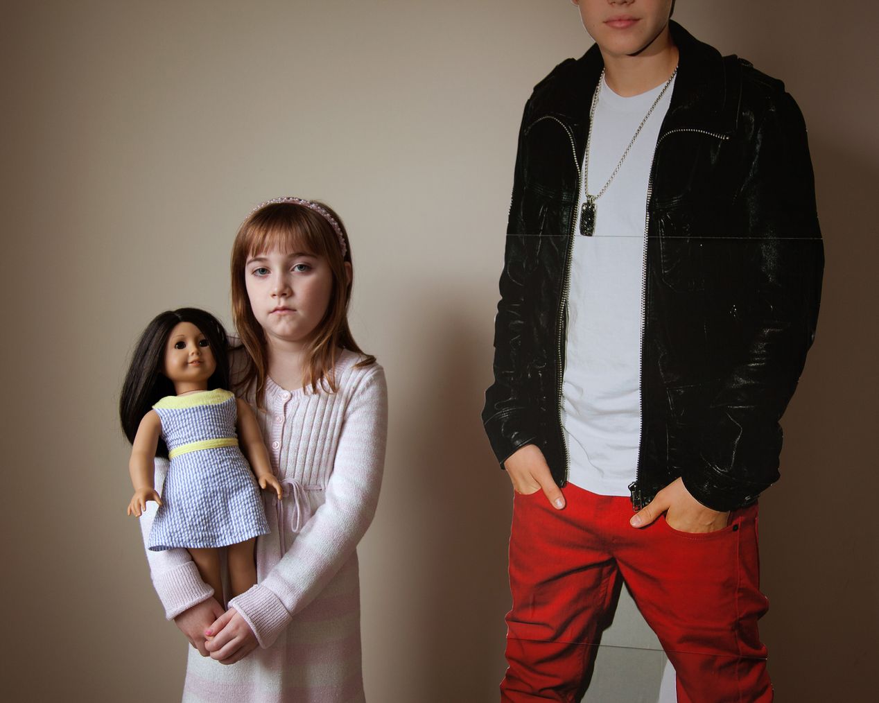 Young girl holding her doll, standing next to a Justin Bieber cutout, environmental portrait photography, Ilona Szwarc, contemporary Los Angeles artist.