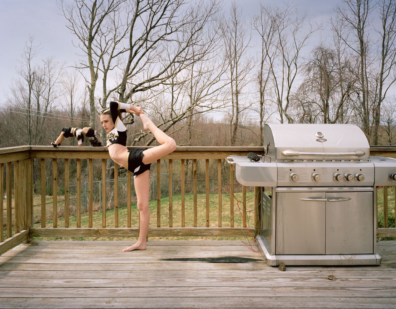 Young athlete and her lookalike doll are posing on a deck next to a large barbeque grill, environmental portrait photography, Ilona Szwarc, contemporary Los Angeles artist.