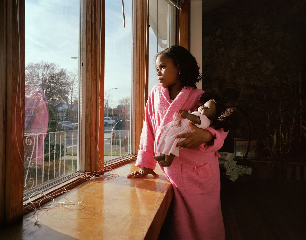 Young girl in a pink robe is holding her lookalike doll and looking out the window, environmental portrait photography, Ilona Szwarc, contemporary Los Angeles artist.