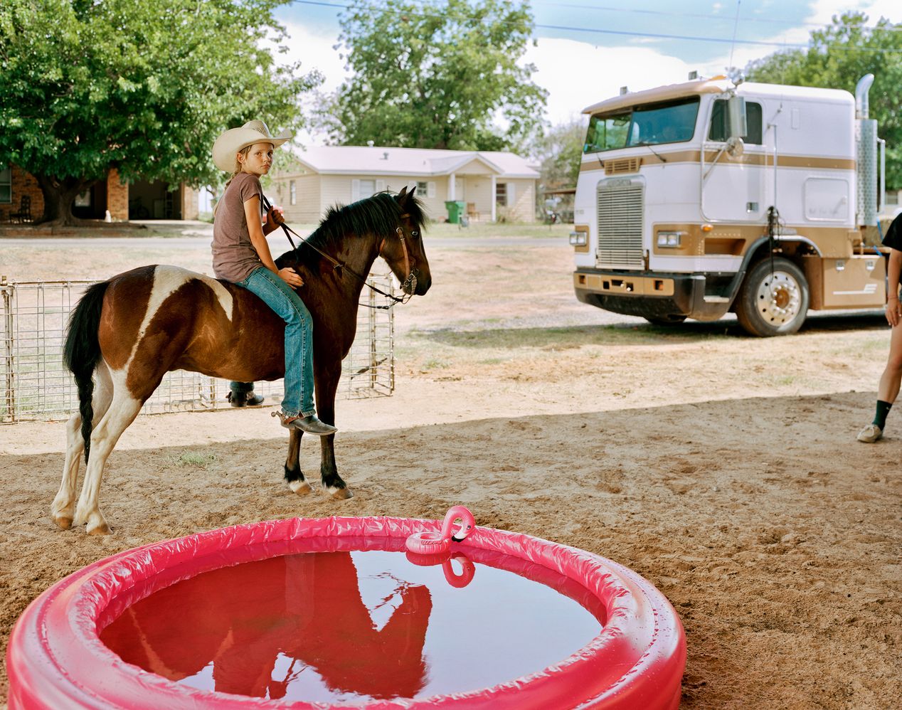 Young cowgirl riding her small horse, next to a truck and an inflatable pink pool, environmental portrait photography, Ilona Szwarc, contemporary Los Angeles artist.